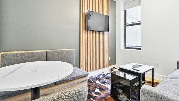 Una habitación moderna con una mesa redonda, sofá gris, paneles de pared de listones de madera, televisor montado y una ventana.