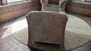 A room with a beige armchair, a glass-top coffee table, wooden panel walls, and large windows with patterned curtains.