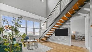A spacious living area with a staircase, a flat-screen TV mounted on the wall, a small round table with a laptop, and a potted plant. - Urban 3-bedroom loft - walk Downtown! (Fort Worth)