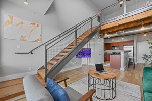 A modern apartment with a staircase, a kitchen, a living area with a sofa and a coffee table, and a TV in the background.