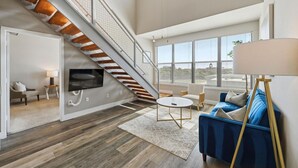 A spacious living area with a staircase, a TV mounted on the wall, a blue sofa, and a large window offering a view. - Large Modern Loft — 3 Bedrooms, walk Downtown! (Fort Worth)