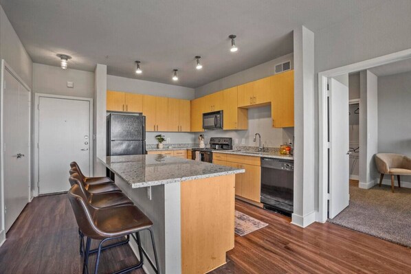 A modern kitchen with a central island, bar stools, wooden cabinets, a refrigerator, microwave, oven, and a dining area with a chair.