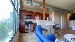 A modern kitchen with stainless steel appliances, a dining area with a white table and two chairs, and a large window offering a city view. - Shop, dine, enjoy - walk Downtown! (Fort Worth)