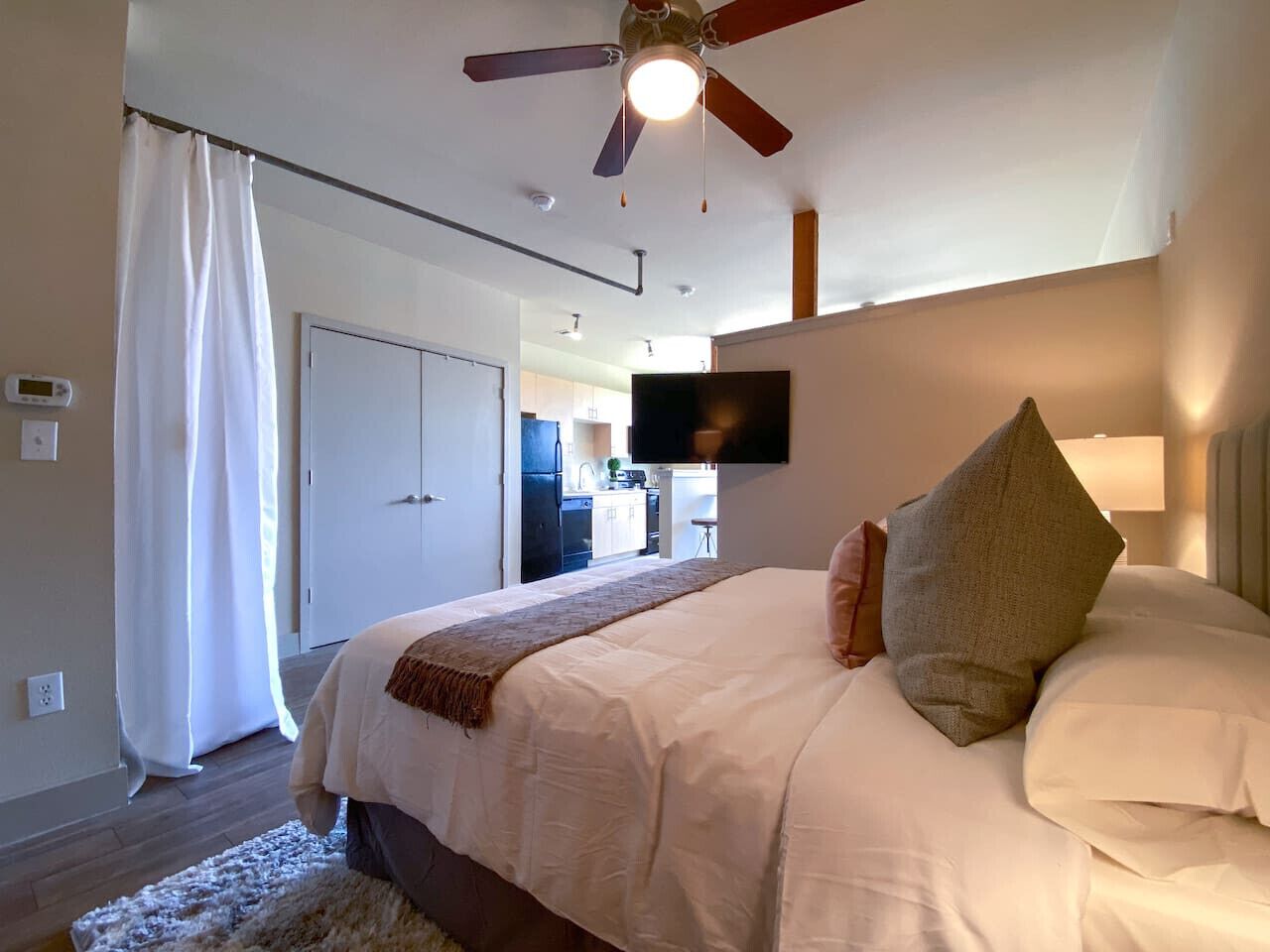 Une chambre à coucher avec un lit, un ventilateur de plafond, une télévision et une vue sur la cuisine.