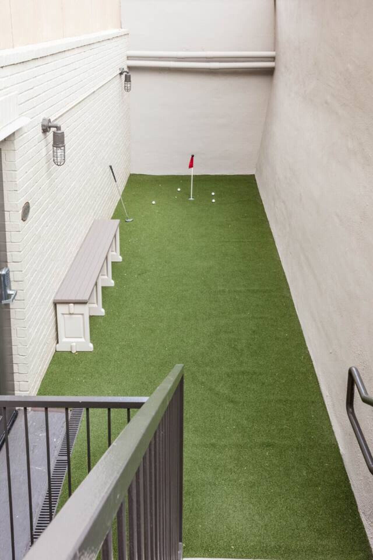 Et smalt indendørs rum med kunstgræs, et rødt flag og en hvid væg med en lysarmatur.