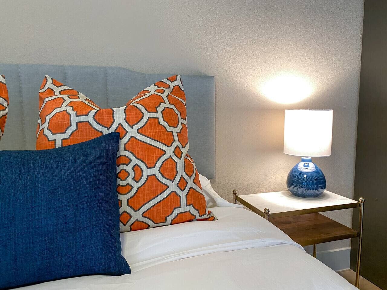 A bed with white bedding, two orange and white patterned pillows, a blue pillow, and a bedside table with a blue lamp.