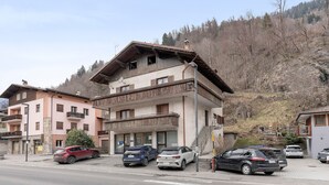 A multi-story building with a balcony, parked cars, and a hilly backdrop. - Room 'Bed and Breakfast Pellizzano' (Pellizzano)