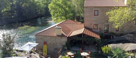 A stone building with a tiled roof, a patio with tables and chairs, and a river in the background.