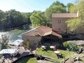 Ein Gebäude aus Stein mit einem Ziegeldach, einer Terrasse mit Tischen und Stühlen und einem Fluss im Hintergrund.