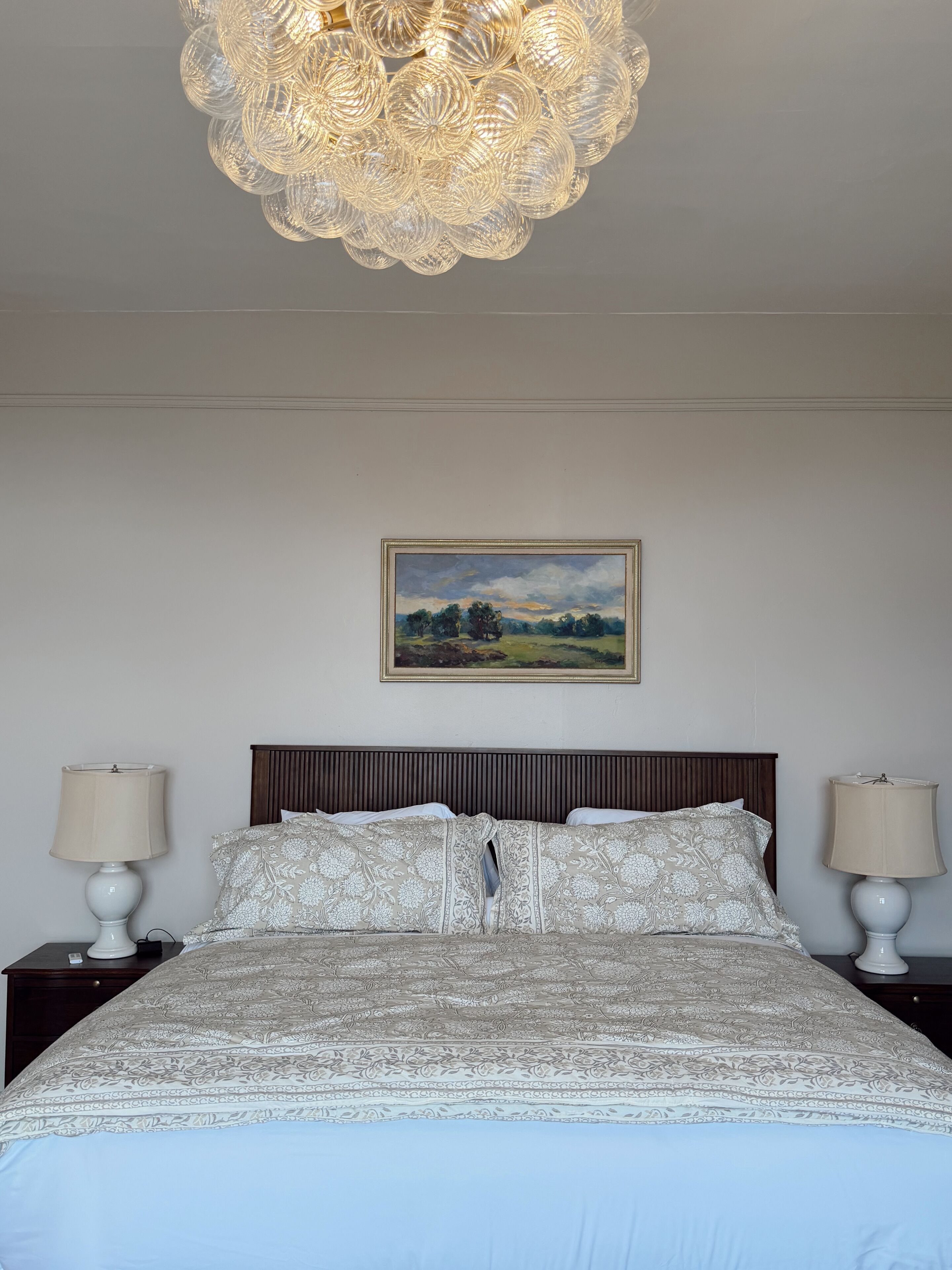 Um quarto com uma cama grande, duas mesas de cabeceira com abajures, um lustre e uma pintura de paisagem na parede.
