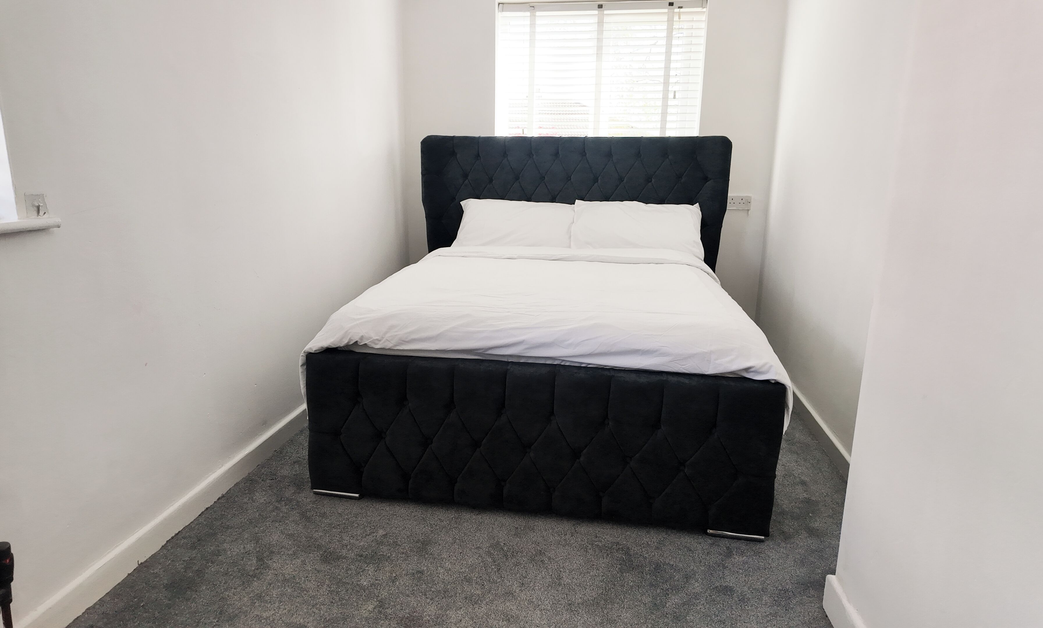 Quarto pequeno e estreito com uma cama de solteiro, roupa de cama branca e um tapete cinzento.
