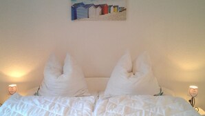 Ein Bett mit weißer Bettwäsche und einer türkisfarbenen Decke, zwei weiße Kissen und ein Bild von Strandhäusern an der Wand.