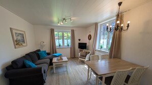 Ein Wohnzimmer mit Sofa, Couchtisch, Esstisch und Stühlen. Es gibt einen Fernseher und eine Deckenleuchte.
