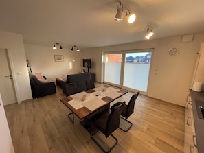 A living room with a sofa, a TV, a dining table with chairs, and a sliding glass door to the outside. - The Wangerkajüte is a modern, cozy first floor vacation apartment with a covered terrace, comfortable furnishings and its own folding boat for excursions on the nearby Wangermeer. (Wangerland)
