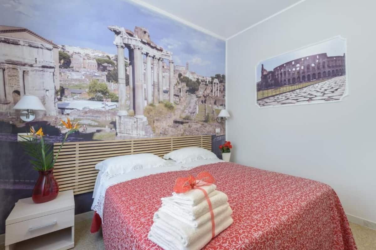 A bedroom with a bed, a nightstand, a lamp, a vase with flowers, and a wall with a mural.