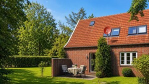 A brick house with a red-tiled roof, a small patio with a table and chairs, and a well-maintained lawn. - The Zugvogel vacation home offers a cozy first floor apartment in a quiet, secluded location near the North Sea - ideal for families and dog owners looking to relax in the fenced garden and natural surroundings. (Wangerland)
