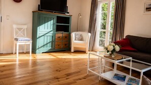 A living room with a television, a green cabinet, a white chair, a brown sofa, and a glass coffee table. - The Zugvogel vacation home offers a cozy first floor apartment in a quiet, secluded location near the North Sea - ideal for families and dog owners looking to relax in the fenced garden and natural surroundings. (Wangerland)