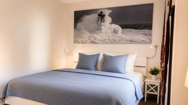 A bedroom with a bed, pillows, a nightstand, a wall-mounted painting of a lighthouse, and a small potted plant. - The Zugvogel vacation home offers a cozy first floor apartment in a quiet, secluded location near the North Sea - ideal for families and dog owners looking to relax in the fenced garden and natural surroundings. (Wangerland)