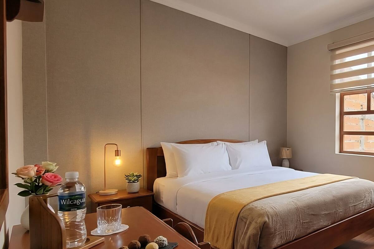 Habitación de hotel con cama, mesita de noche, lámpara, ventana con persianas y cartel de bienvenida.