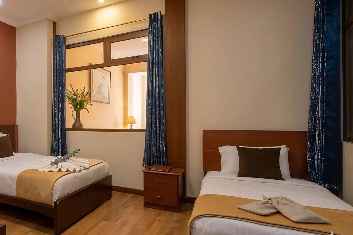 Habitación de hotel con dos camas, una ventana con cortinas, un cuadro en la pared y una bandeja con una taza y galletas.