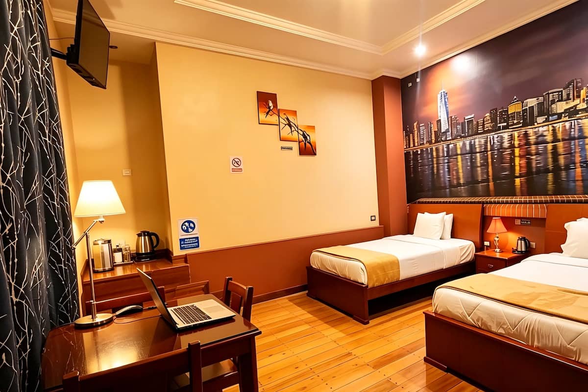 Habitación de hotel con dos camas, un escritorio con una computadora portátil, cafetera y un cuadro de un paisaje urbano.