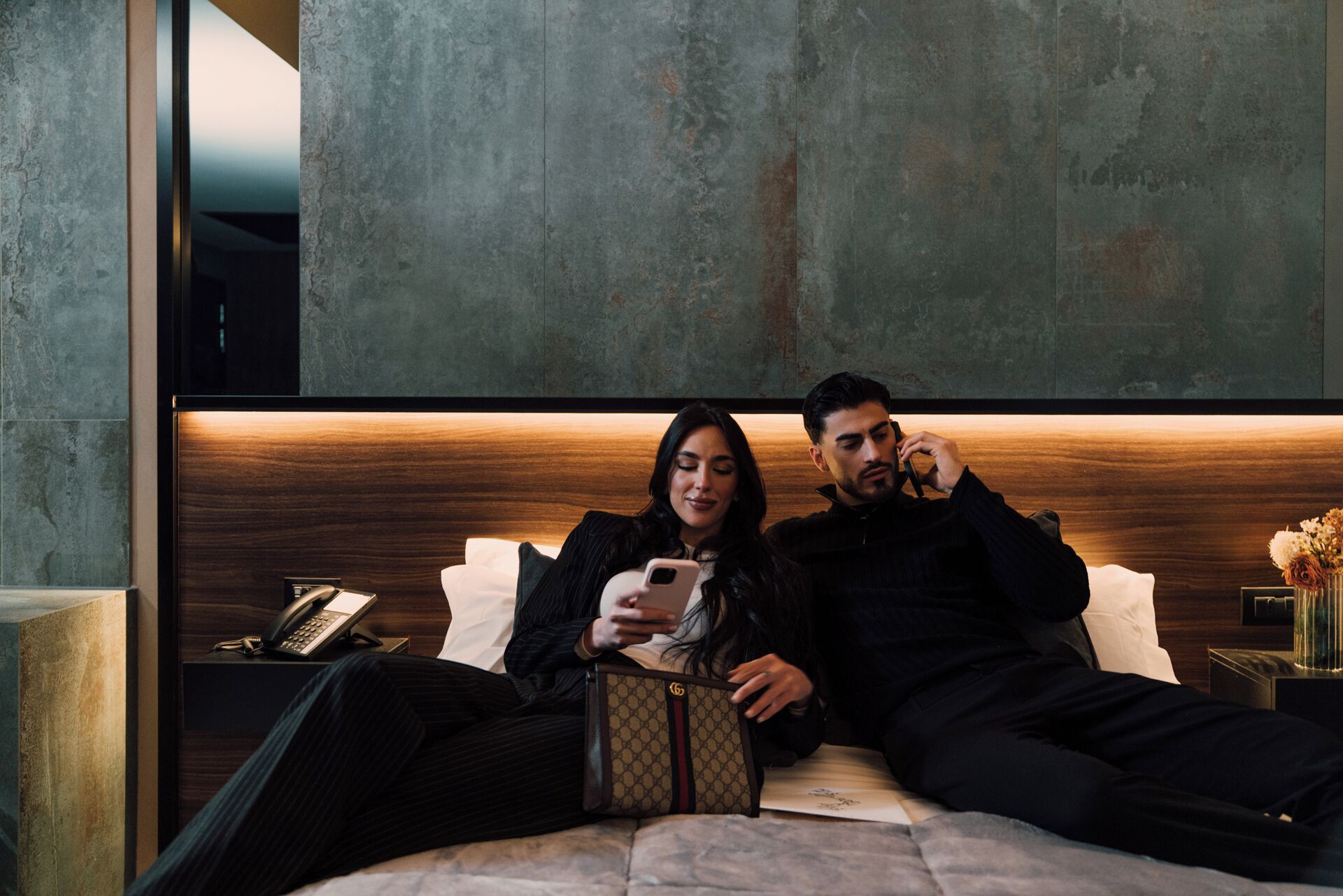 Een moderne slaapkamer met een houten hoofdbord, een nachtkastje met een lamp en een bijzettafeltje met een telefoon en sleutels.