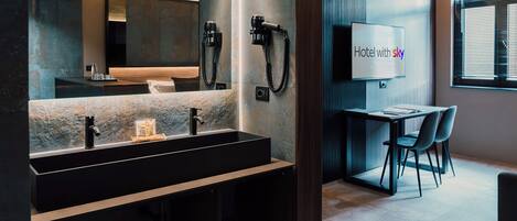 Un baño moderno con un lavamanos grande, dos secadores de pelo y un espejo. Hay una mesita con una silla y un letrero que dice 'Hotel con cielo'.