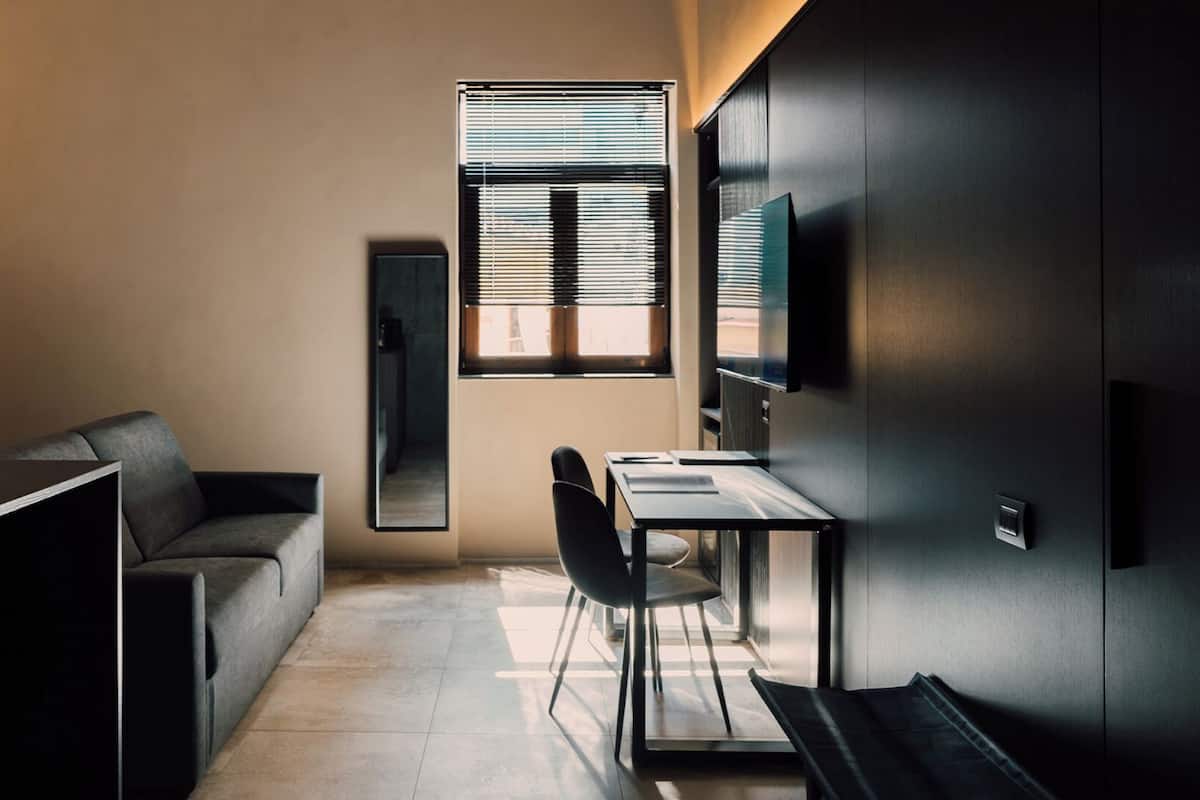 Quarto compacto com sofá, escrivaninha, cadeira, televisão e janela com persianas.