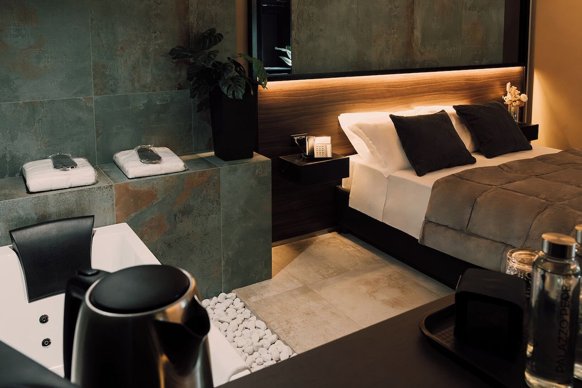 Quarto de hotel moderno com cama, mesa de cabeceira, poltrona e área de estar com mesa.