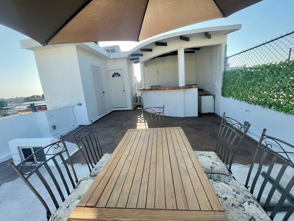 A covered outdoor seating area with a wooden table and metal chairs, a white door, and a view of the cityscape.