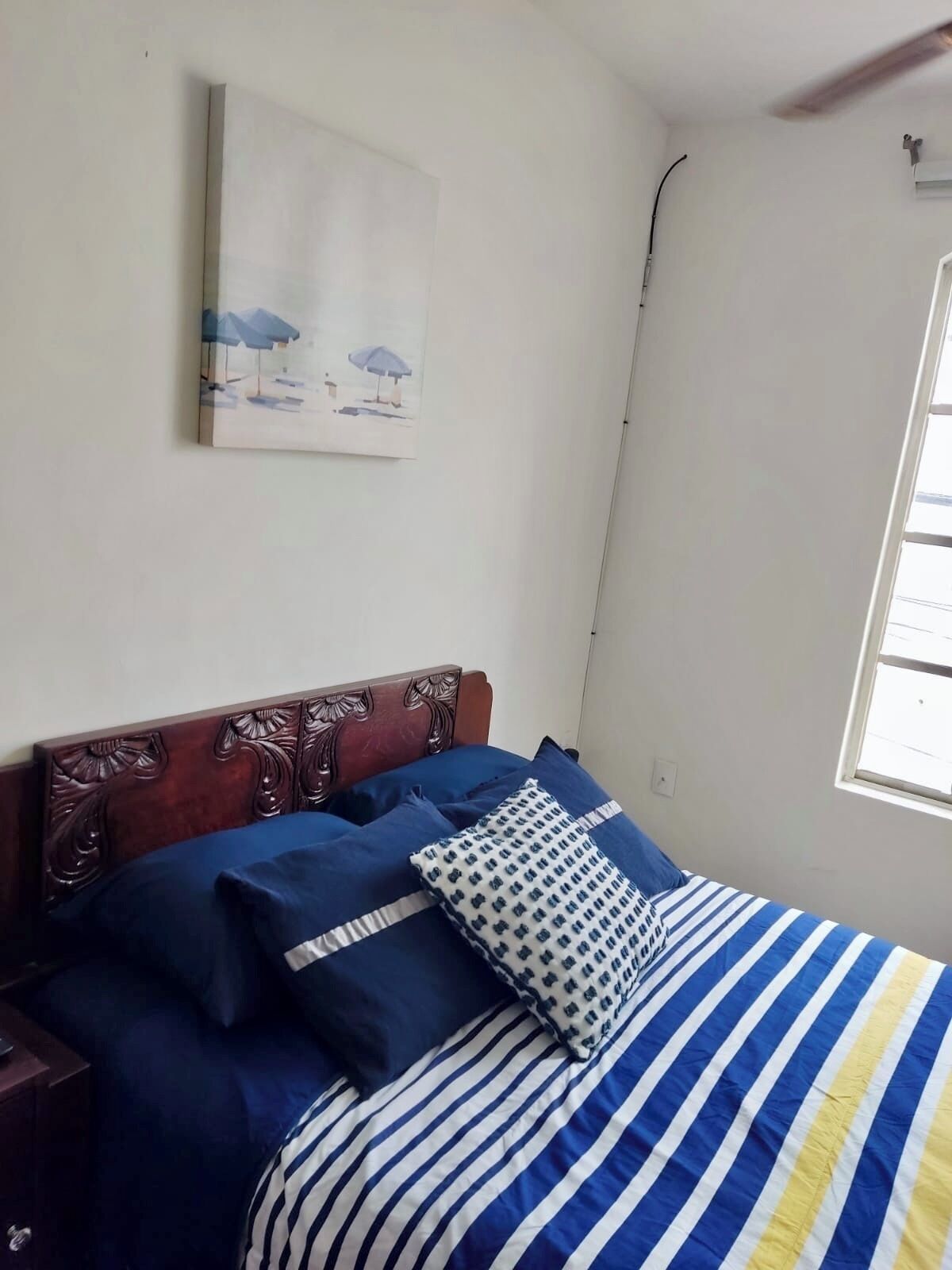 A bedroom with a wooden headboard, a bed with striped bedding, a blue pillow, and a painting on the wall.