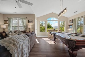 Ein Zimmer mit Poolbillard, einer Couch mit Decke, einem Deckenventilator und großen Fenstern mit Vorhängen.