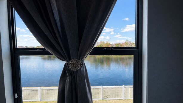 Ett fönster med en gardin som är knuten i mitten, med utsikt över en vattenyta och ett vitt staket.