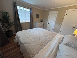 A bedroom with a bed, a dresser, a door, a window with curtains, and a potted plant.