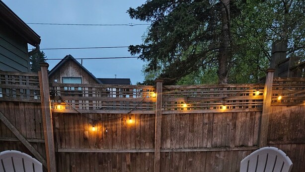 A square fire pit table with two Adirondack chairs, surrounded by a wooden fence and string lights. - Downtown home w/patio in historic Pilot's Row (Anchorage)