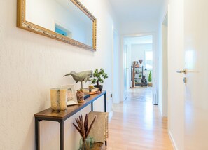 A hallway with a console table displaying decorative items, a large mirror, and a wooden floor leading to a room with a Buddha statue and a bookshelf.