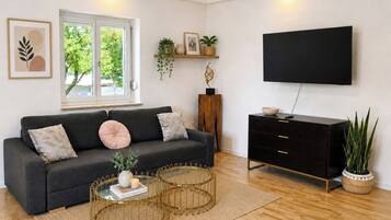 Ein modernes Wohnzimmer mit einem dunkelgrauen Sofa, runden Couchtischen, einem schwarzen Fernseher an der Wand und Holzböden.