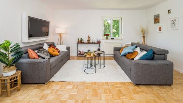 Een moderne woonkamer met twee grijze banken, een witte vloerkleed, een houten vloer, een aan de muur bevestigde televisie, een raam met uitzicht op bomen en een tafeltje met decoratieve objecten.