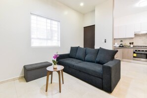 Un salon moderne avec un canapé gris, une petite table ronde sur laquelle est posée une orchidée en pot, et un coin cuisine à l’arrière-plan.