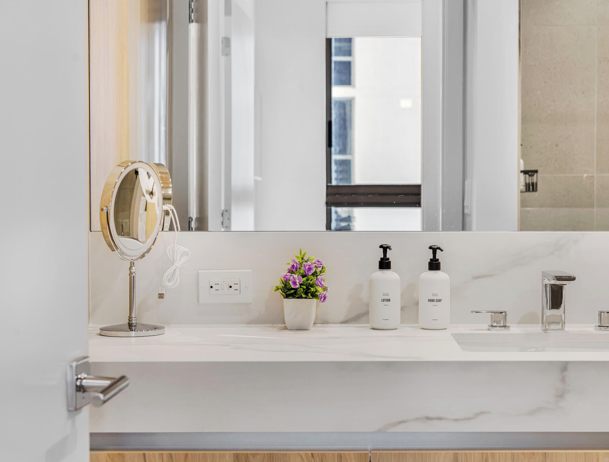 A bathroom with a white marble countertop, a round mirror, a potted plant, and two bottles of lotion and hand soap.