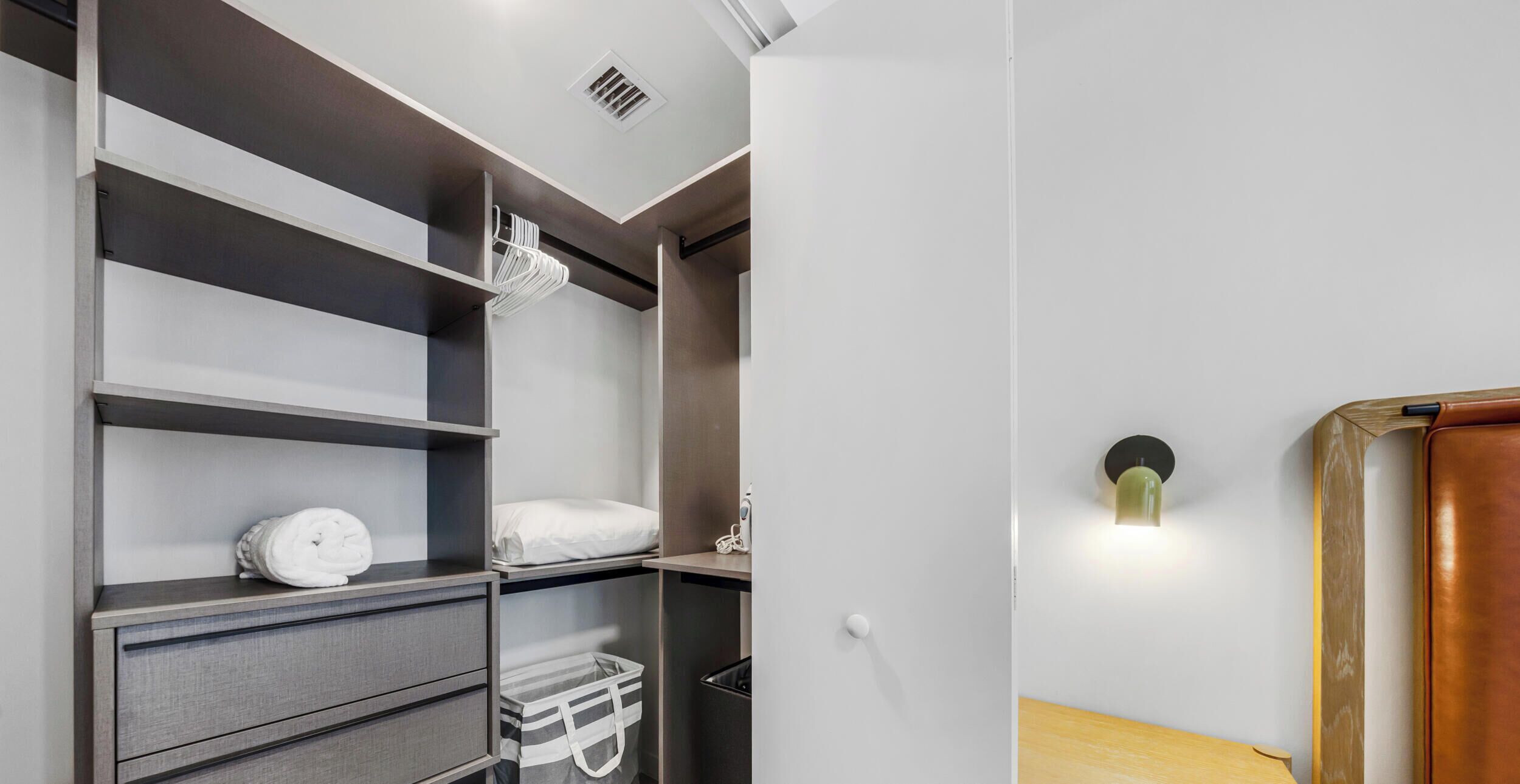 Une chambre d’hôtel moderne dotée d’un placard intégré, d’une table de chevet et d’un luminaire fixé au mur.
