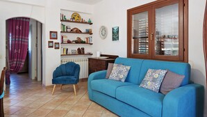 A living room with a blue sofa, a wooden cabinet with glass doors, and a dining area with a chair and table. - Villa 'Diego' with Sea View, Wi-Fi and Air Conditioning (Favignana)