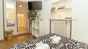 A modern hotel room with a bed, a TV, a radiator, a shelf with books, and a small plant. - riMINI house - One Bedroom Apartment, Sleeps 2 (Rimini)