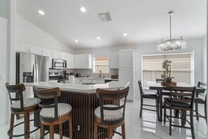 A modern kitchen with white cabinetry, a central island with bar stools, and a dining area with a chandelier. - 5-Bed Villa w/ Private Pool (Davenport)