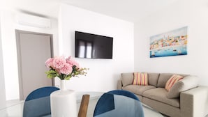 Ein modernes Wohnzimmer mit einem Glastisch, blauen Stühlen, einem beigen Sofa, einem Flachbildfernseher und einem Gemälde mit einer Küstenlandschaft.