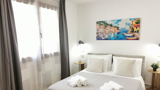 Ein ordentlich gemachtes Bett mit Kissen und gerollten Handtüchern, ein Gemälde mit Meeresblick und ein Fenster mit Vorhängen.