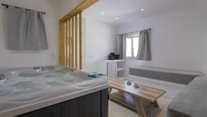 Une chambre moderne dotée d’un jacuzzi, de poutres en bois, d’un canapé, d’une petite table et d’une fenêtre avec des rideaux.