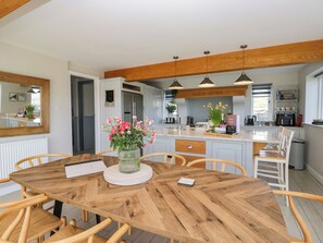 A modern kitchen with a central island, wooden ceiling beams, pendant lighting, and a dining area with a wooden table and chairs.