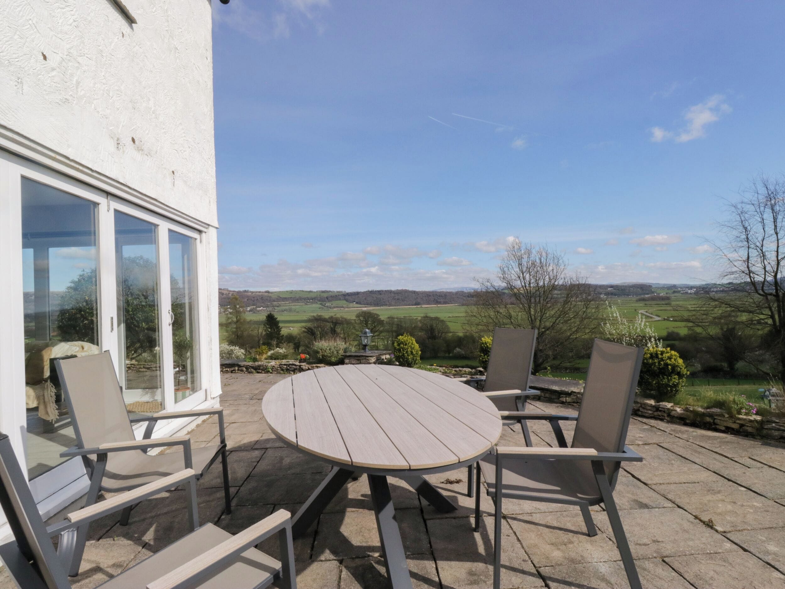 An outdoor patio with a round table and chairs, a white building with glass doors, and a view of green fields and hills.