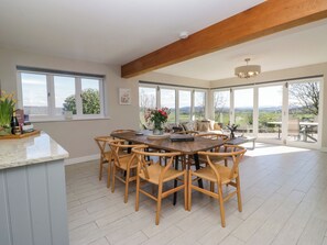 A spacious dining area with a wooden table and chairs, large windows with a view of the countryside, and a wooden ceiling beam. - Barn Howe (Kendal)
